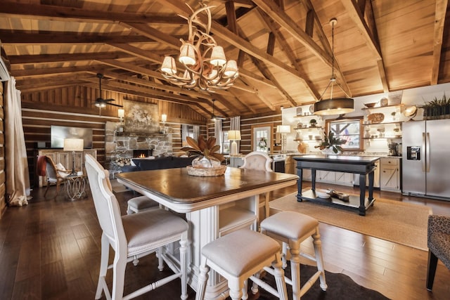 dining room with dark wood-type flooring, a chandelier, lofted ceiling with beams, a stone fireplace, and wooden ceiling