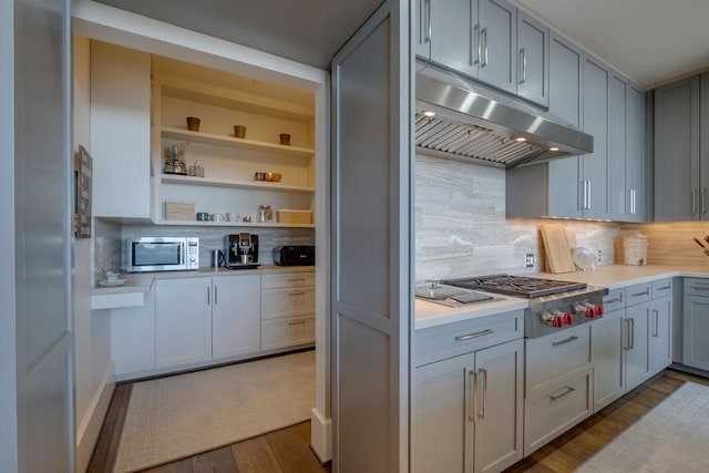 kitchen with under cabinet range hood, light countertops, appliances with stainless steel finishes, wood finished floors, and open shelves