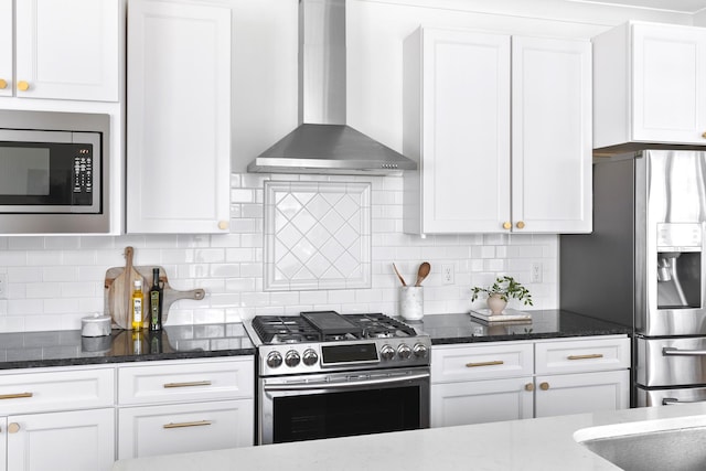 kitchen featuring white cabinets, stainless steel appliances, wall chimney range hood, and dark stone countertops