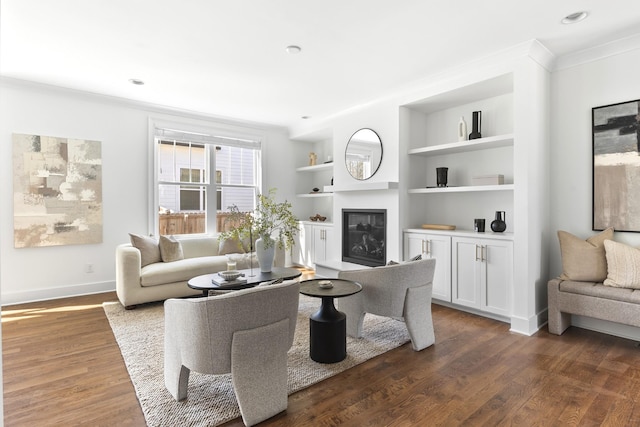 living room with built in features, dark wood-style floors, baseboards, ornamental molding, and a glass covered fireplace