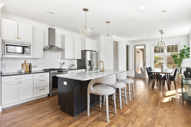 kitchen with dark wood-type flooring, wall chimney range hood, an island with sink, decorative backsplash, and stainless steel appliances