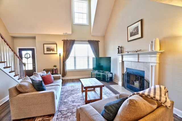 living room featuring high vaulted ceiling, a tiled fireplace, wood finished floors, stairway, and baseboards