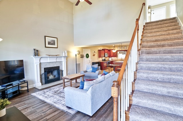 living area with dark wood-type flooring, a towering ceiling, a fireplace, ceiling fan, and stairs