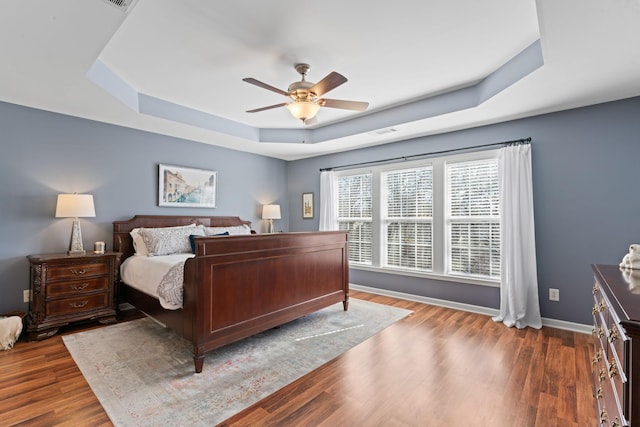 bedroom featuring wood finished floors, visible vents, baseboards, a tray ceiling, and ceiling fan