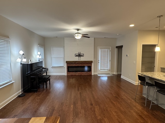 living area featuring a brick fireplace, baseboards, dark wood-type flooring, and a ceiling fan