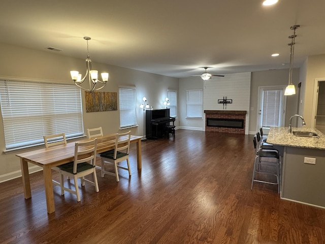 dining space with visible vents, ceiling fan with notable chandelier, dark wood-style floors, baseboards, and a brick fireplace