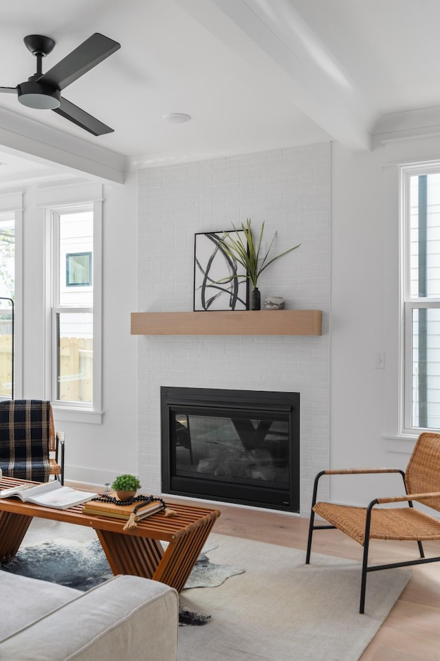 living room featuring a ceiling fan, beam ceiling, a brick fireplace, and wood finished floors