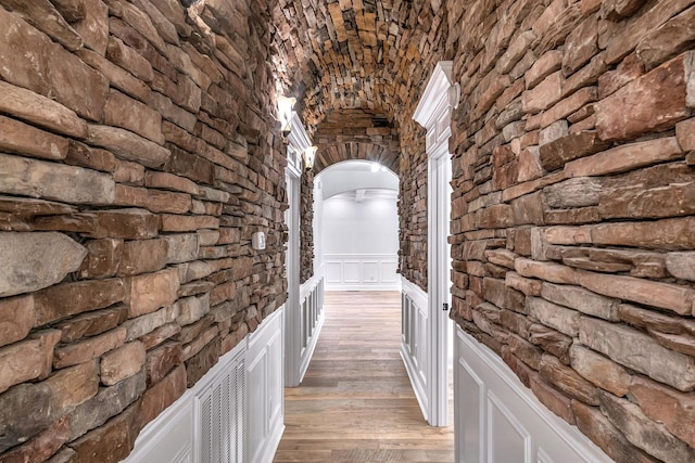 hallway with a wainscoted wall, arched walkways, brick ceiling, light wood finished floors, and lofted ceiling