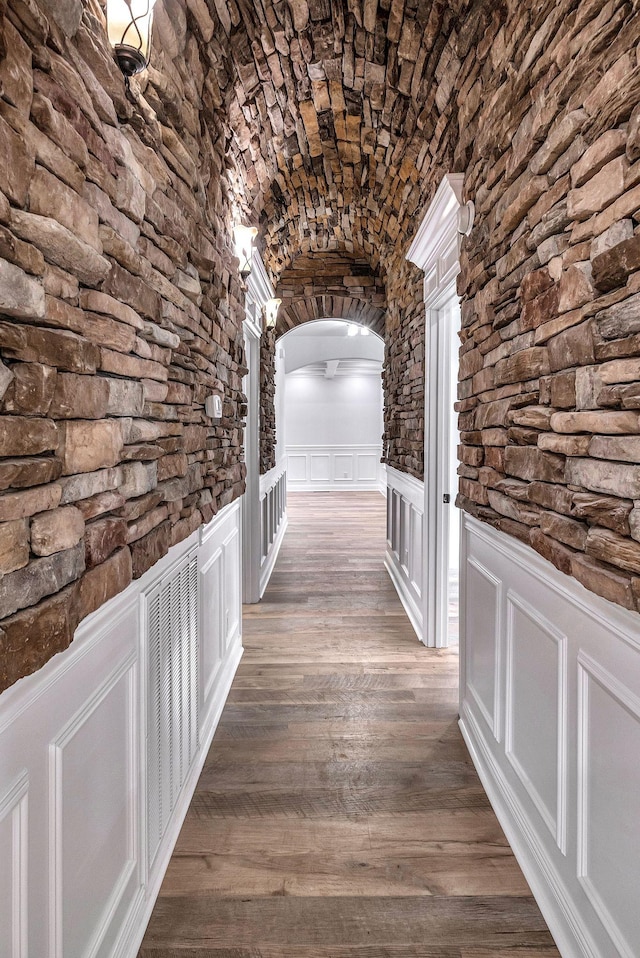 hallway with arched walkways, dark wood-style flooring, vaulted ceiling, a decorative wall, and brick ceiling