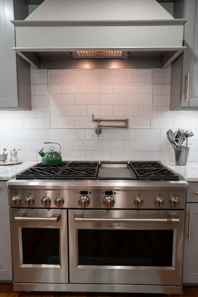 kitchen with backsplash, light stone countertops, custom range hood, range with two ovens, and gray cabinets