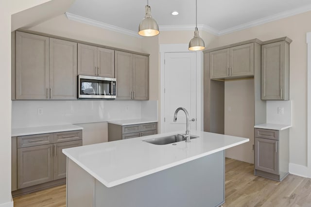 kitchen featuring light wood finished floors, stainless steel microwave, crown molding, a center island with sink, and a sink