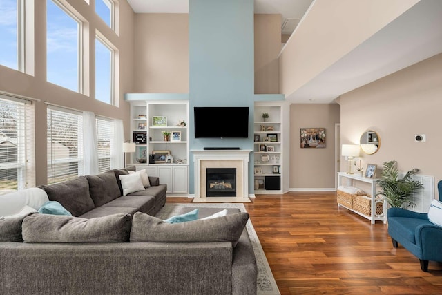 living room with a fireplace with flush hearth, built in features, wood finished floors, baseboards, and a towering ceiling