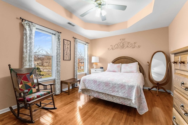 bedroom with visible vents, light wood-style flooring, a tray ceiling, baseboards, and ceiling fan