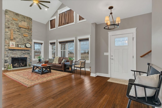 foyer with high vaulted ceiling, ceiling fan with notable chandelier, dark wood-style floors, a stone fireplace, and baseboards