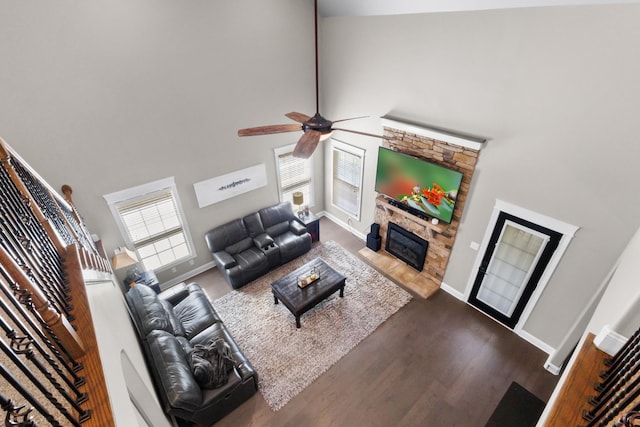 living area featuring dark wood-style floors, a ceiling fan, baseboards, a stone fireplace, and a towering ceiling