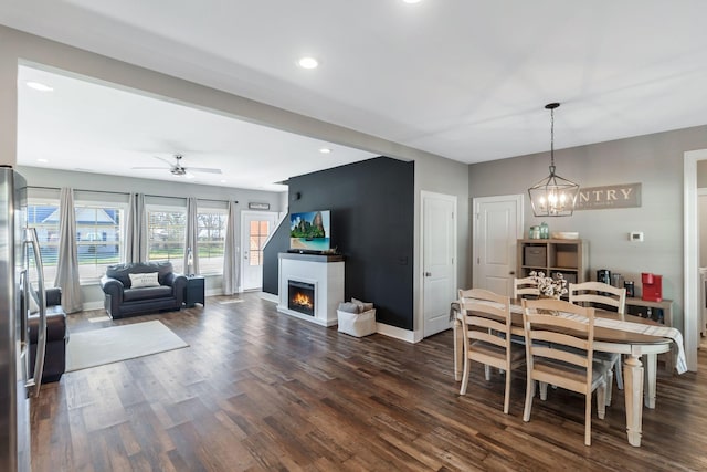 dining area with baseboards, dark wood finished floors, recessed lighting, a lit fireplace, and ceiling fan with notable chandelier