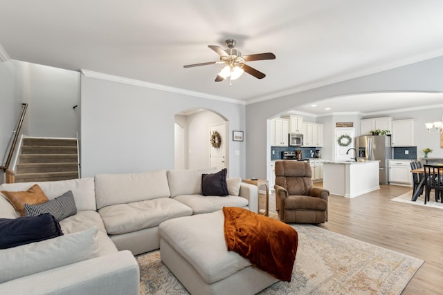 living room with ceiling fan with notable chandelier, arched walkways, crown molding, light wood finished floors, and stairs