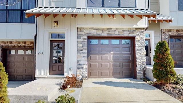 doorway to property with metal roof, a garage, stone siding, driveway, and a standing seam roof