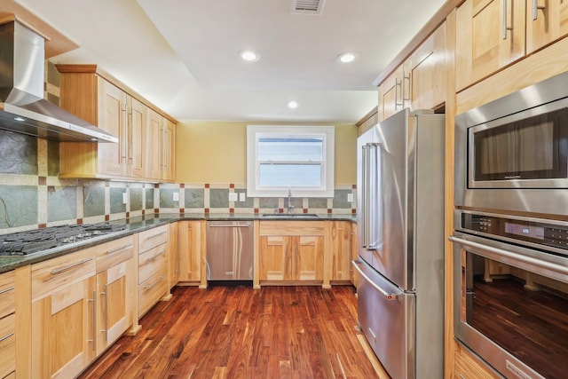 kitchen featuring light brown cabinets, dark wood-style flooring, a sink, appliances with stainless steel finishes, and wall chimney range hood