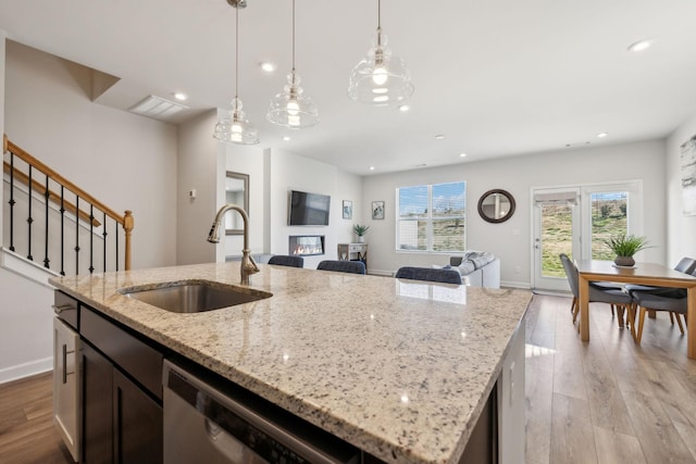 kitchen with light wood finished floors, a sink, an island with sink, a glass covered fireplace, and stainless steel dishwasher