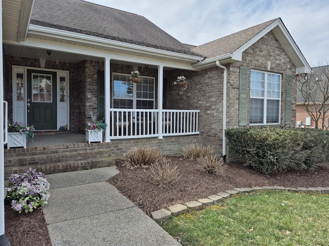 doorway to property with brick siding, a porch, and a shingled roof