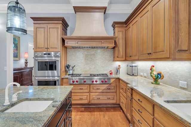 kitchen featuring light wood finished floors, premium range hood, ornamental molding, appliances with stainless steel finishes, and a sink