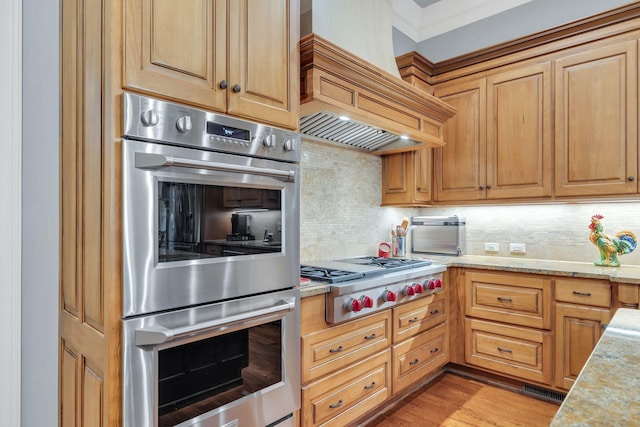 kitchen with backsplash, custom range hood, light stone counters, light wood-style flooring, and appliances with stainless steel finishes