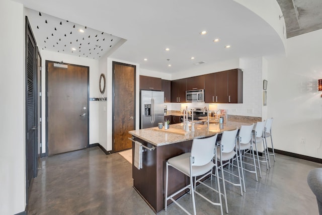 kitchen with finished concrete flooring, stainless steel appliances, dark brown cabinetry, a peninsula, and a breakfast bar area