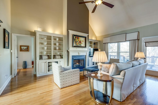 living area featuring light wood finished floors, baseboards, a glass covered fireplace, high vaulted ceiling, and a ceiling fan