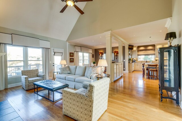 living area featuring a ceiling fan, baseboards, high vaulted ceiling, light wood-style flooring, and decorative columns