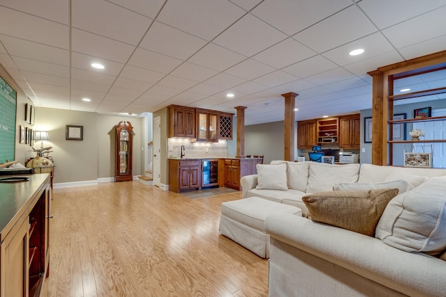 living room with light wood-type flooring, beverage cooler, baseboards, decorative columns, and wet bar