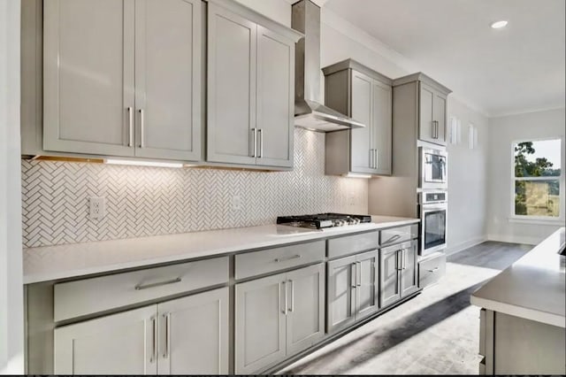 kitchen featuring gray cabinets, backsplash, crown molding, wall chimney range hood, and stainless steel oven