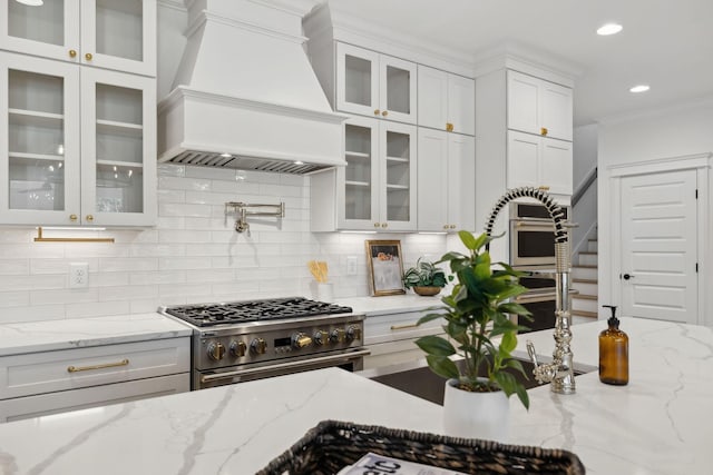 kitchen featuring custom exhaust hood, glass insert cabinets, high end stainless steel range, white cabinetry, and tasteful backsplash