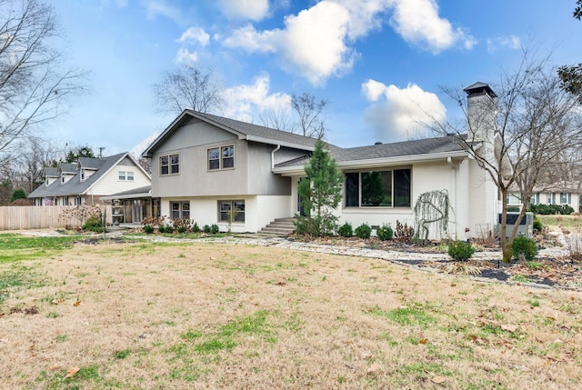 split level home with stucco siding, a chimney, a front lawn, and fence