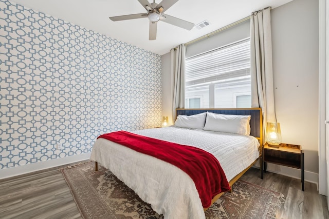 bedroom featuring wood finished floors, baseboards, visible vents, wallpapered walls, and an accent wall