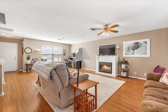 living room with a ceiling fan, baseboards, light wood finished floors, a textured ceiling, and a glass covered fireplace