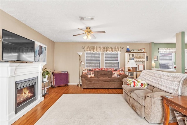 living area with visible vents, a ceiling fan, a textured ceiling, a glass covered fireplace, and wood finished floors