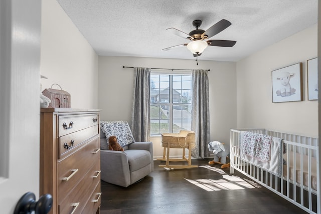 bedroom with a crib, dark wood-type flooring, a ceiling fan, and a textured ceiling