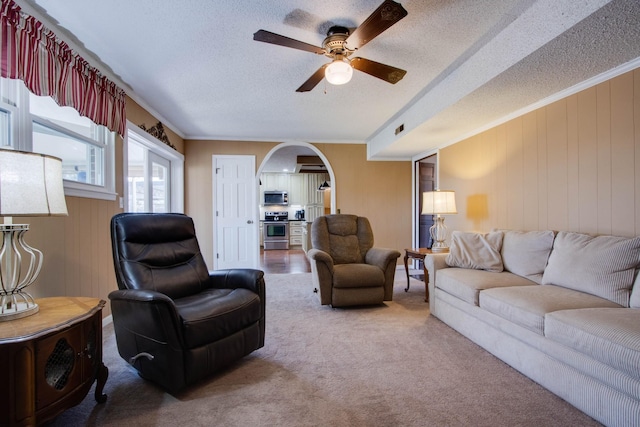 carpeted living area with a textured ceiling, a ceiling fan, arched walkways, and ornamental molding