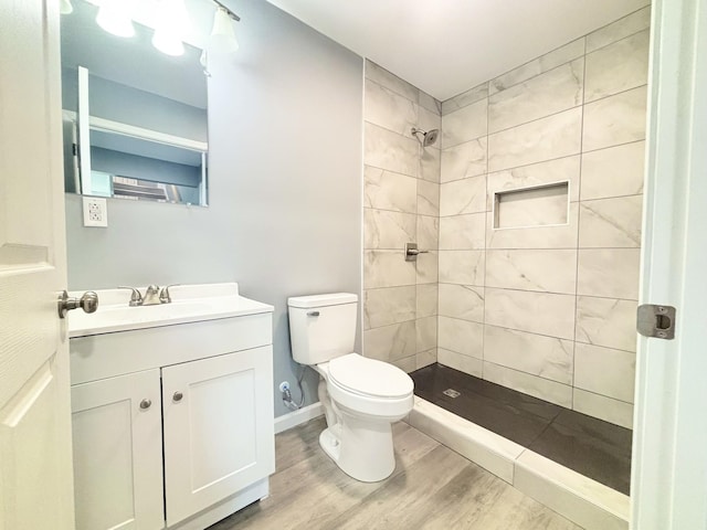 bathroom featuring toilet, wood finished floors, tiled shower, baseboards, and vanity