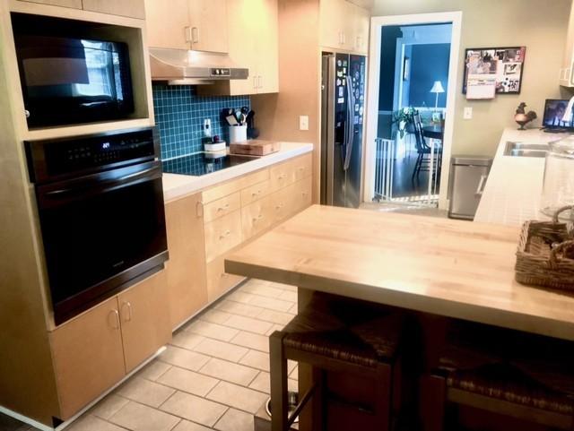 kitchen with light tile patterned floors, black appliances, light countertops, under cabinet range hood, and tasteful backsplash