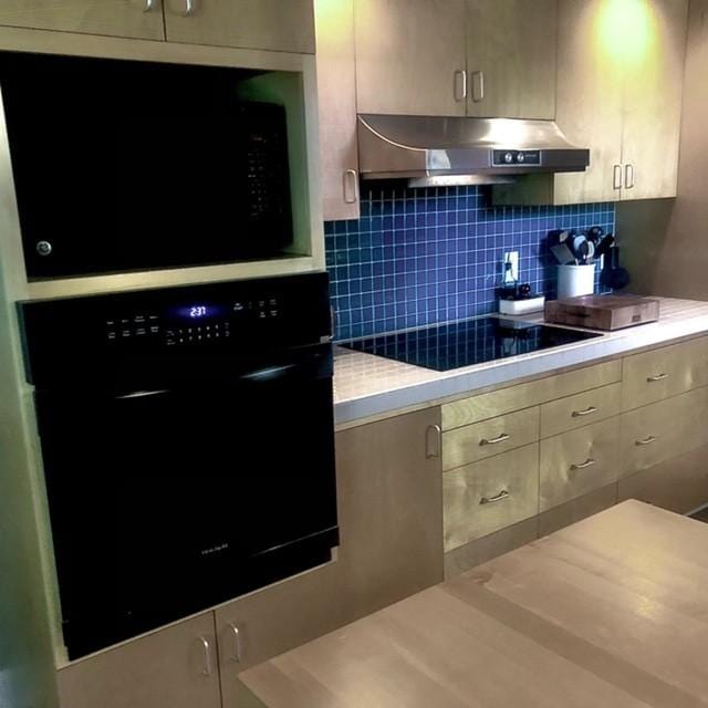 kitchen featuring under cabinet range hood, decorative backsplash, black appliances, and light countertops