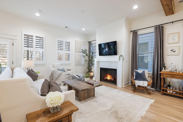 living area with light wood-style flooring, recessed lighting, a lit fireplace, ornamental molding, and beamed ceiling