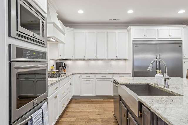 kitchen with light wood-type flooring, custom range hood, built in appliances, white cabinetry, and tasteful backsplash