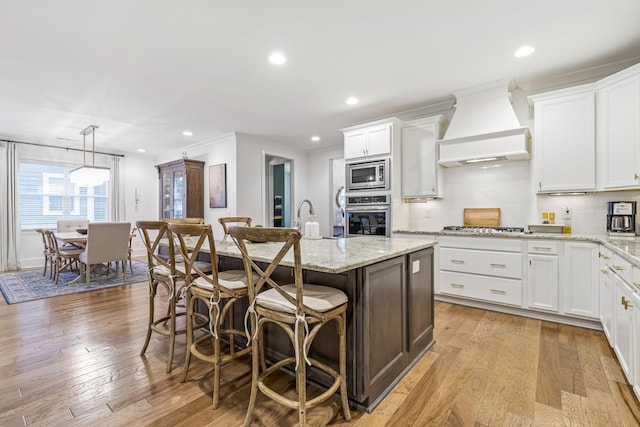 kitchen with light wood finished floors, a kitchen bar, an island with sink, custom range hood, and appliances with stainless steel finishes