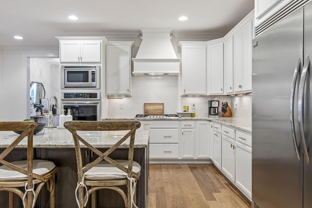 kitchen featuring wood finished floors, premium range hood, built in appliances, white cabinetry, and backsplash