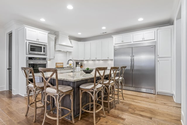 kitchen featuring light stone counters, premium range hood, light wood-style flooring, built in appliances, and white cabinetry