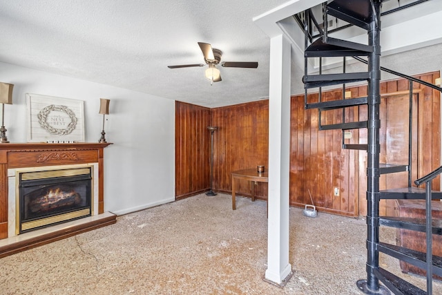 unfurnished living room featuring wooden walls, a ceiling fan, stairs, a textured ceiling, and a glass covered fireplace