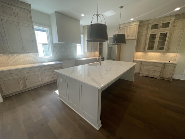 kitchen with tasteful backsplash, a sink, glass insert cabinets, a center island with sink, and dark wood-style flooring