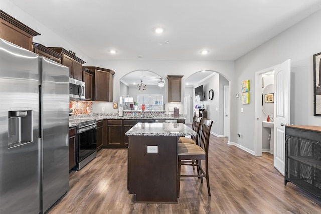 kitchen featuring a kitchen island, arched walkways, dark brown cabinetry, dark wood-type flooring, and appliances with stainless steel finishes